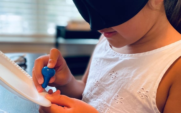 Picture of a blindfolded, young girl, being taught to use a stylus.